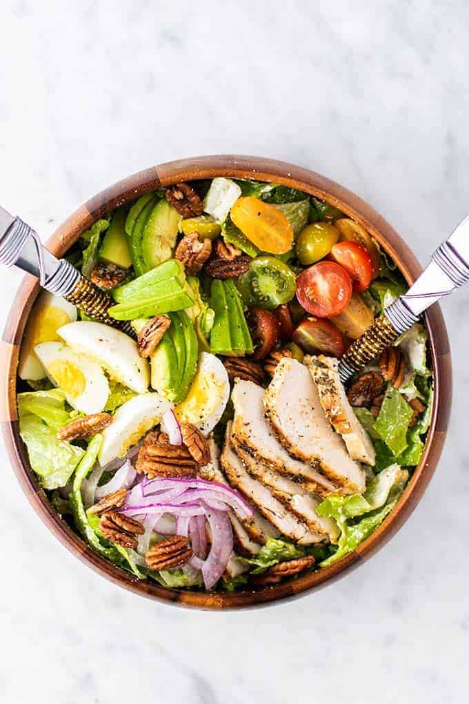 Chicken salad in a wooden bowl with avocados, eggs, red onion, tomatoes, pecans, chicken, and lettuce.